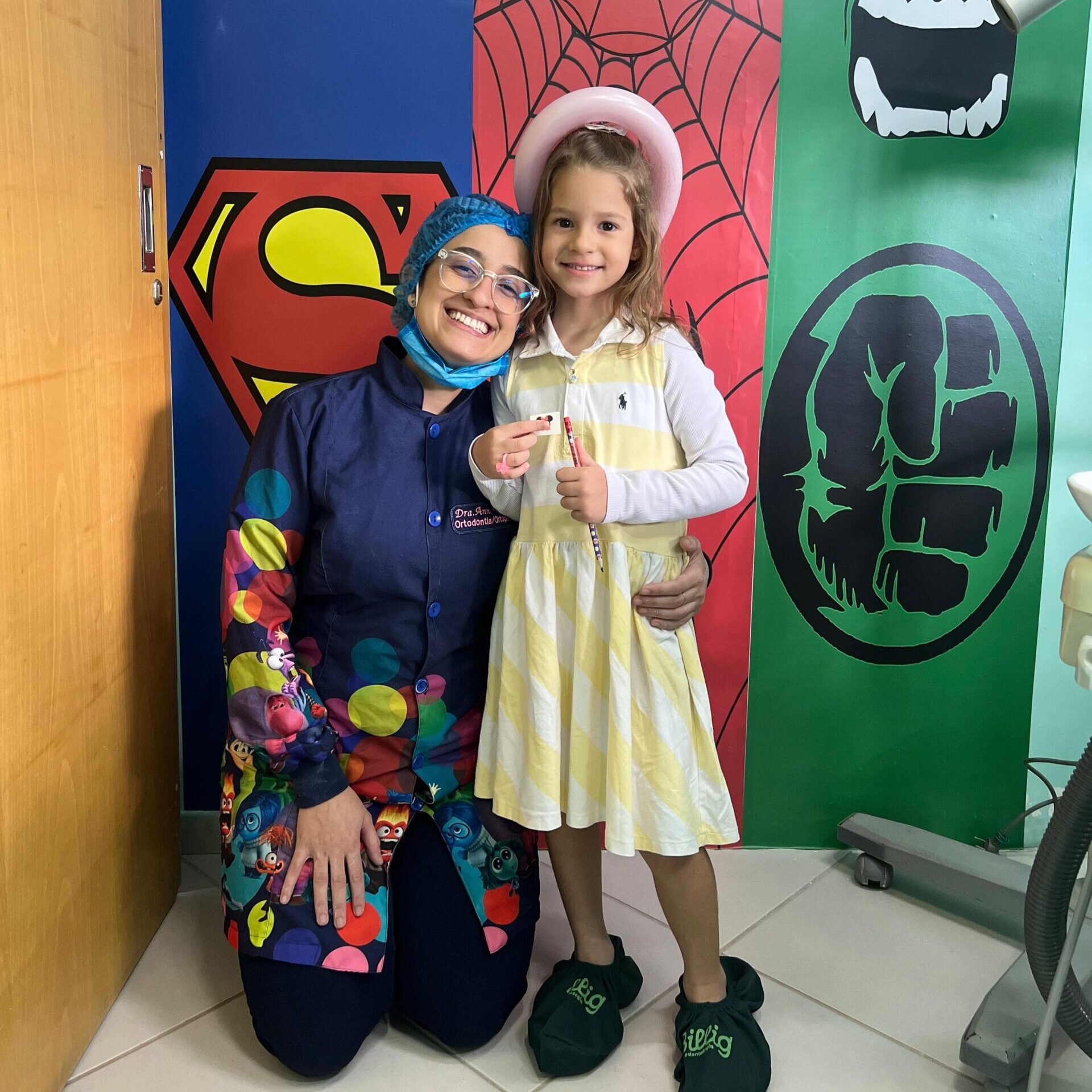 Ortodontista posando com paciente infantil em consultório decorado com temática de super-heróis durante tratamento ortodôntico